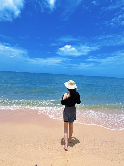 A woman wearing a sun hat, black top, and gray skirt stands barefoot on a sandy beach looking out at a calm blue ocean under a bright blue sky with scattered clouds.