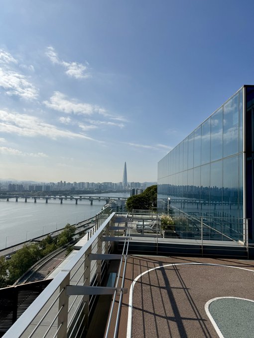 View from a rooftop terrace overlooking a river with bridges, a modern glass building on the right, and a tall skyscraper visible in the distance under a partly cloudy blue sky.