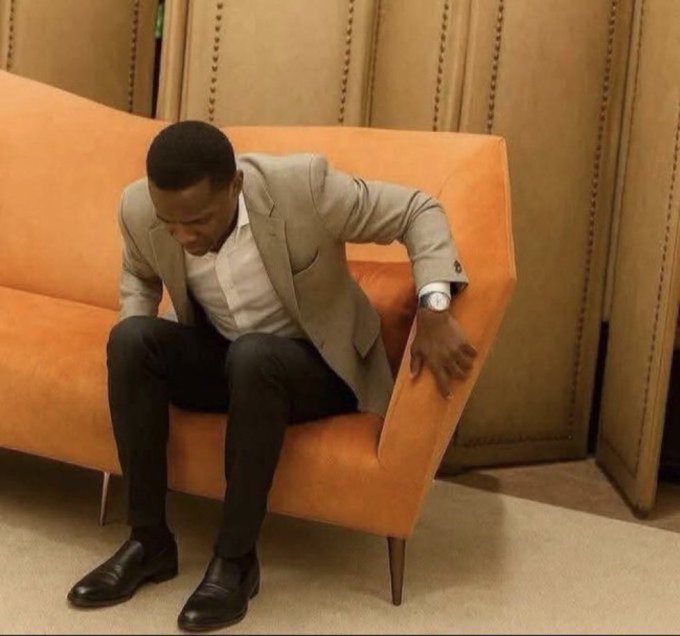 The image features a man sitting on an orange couch, wearing a suit and tie. He has his hand resting on the arm of the couch, giving off a relaxed yet professional vibe. The background includes a wall with a clock mounted on it, suggesting that time is of importance in this setting. The caption "GN CT Don't forget to talk about @quipnetwork daily" implies that the image could be related to a social media platform or blog where the man's outfit and style are being discussed.
