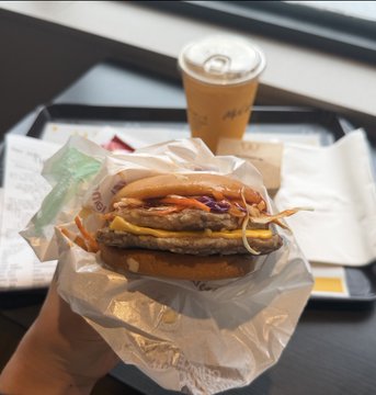 The image shows a hand holding up a sandwich wrapped in plastic, with a drink placed next to it. The sandwich appears to be a burger or a similar type of food item. The drink has a straw in it, which suggests that it might be a soft drink like soda or juice. The scene takes place on a dining table, and the person holding up the sandwich is likely preparing for a meal or enjoying their lunch.