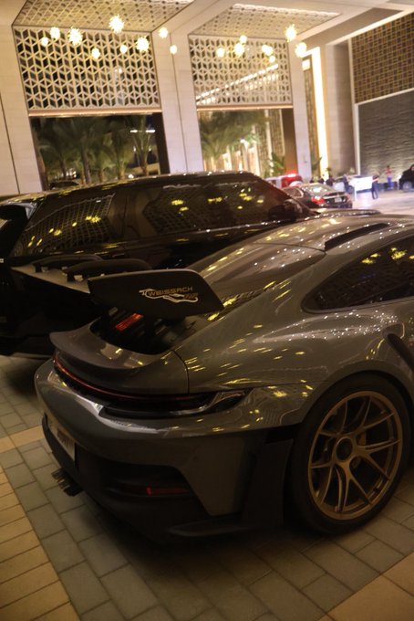 The rear view of a grey Porsche 911 GT3 sports car with a large rear wing parked outside a building at night, featuring bronze-colored wheels.