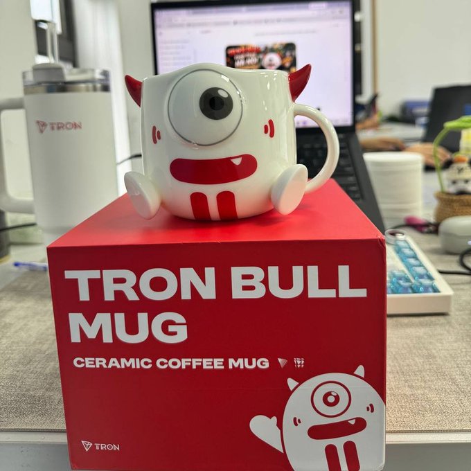 A 'TRON BULL MUG' ceramic coffee mug shaped like a cute monster character with one large eye and red horns, displayed on top of its red product packaging box on a desk with a laptop in the background.
