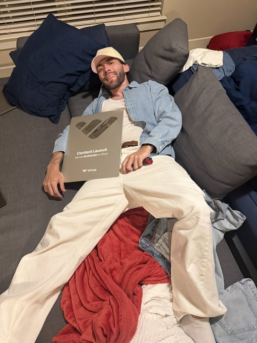 A man in a denim shirt and white pants lies sprawled across a gray sectional surrounded by throw pillows, smiling as he holds a silver "Content Launch" plaque from Whop. The celebratory image reflects his milestone of scaling from $0 to $1M in 3 months and now $1M/month within 6 months, marking his next goal of multi-million monthly earnings.