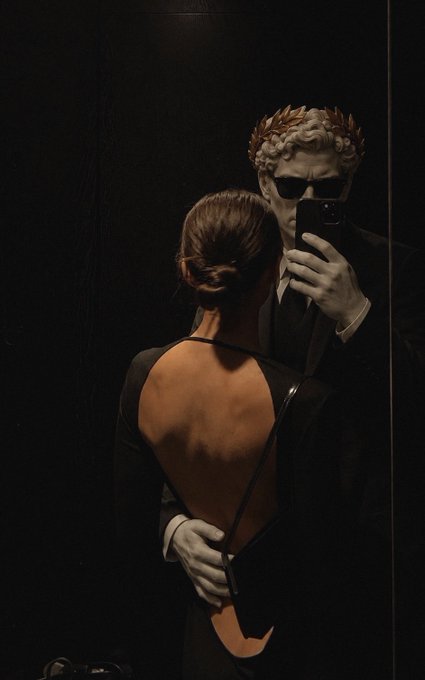 A dimly lit mirror selfie featuring a person in a backless black dress and a striking sculptural figure wearing a laurel wreath and dark sunglasses, holding a smartphone to take the photo.