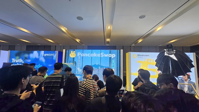 A crowded cryptocurrency conference with attendees, featuring prominent branding for PancakeSwap, Bitcoin.com, and Thailand at booths with large screens.