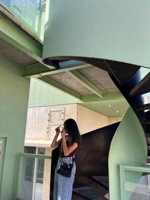 A young woman with long dark hair takes a mirror selfie with a camera, standing near a striking mint-green spiral staircase in a modern architectural space with concrete beams and beige walls. Her outfit—a crop top and denim jeans with a small black crossbody bag—complements the minimalist aesthetic, while the vivid green background plays on her caption's wish for "green candles" in trading.
