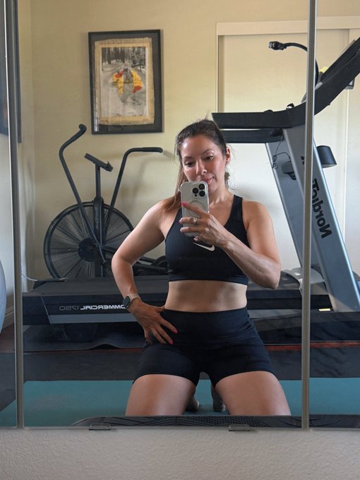A woman in black athletic wear takes a mirror selfie in a home gym with a treadmill and elliptical machine in the background.