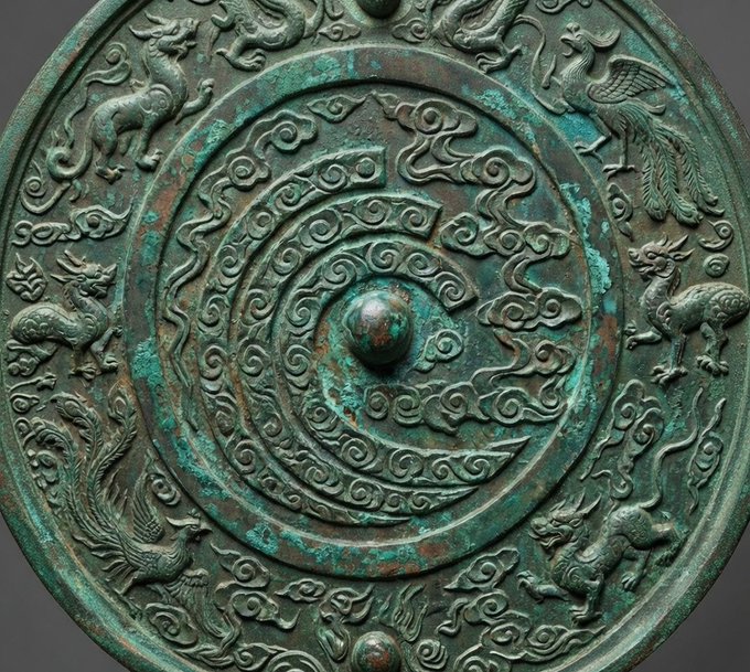 An ancient bronze mirror with green patina, featuring intricate swirling designs and animal figures (dragon, phoenix, and others) around its perimeter and concentric circles.