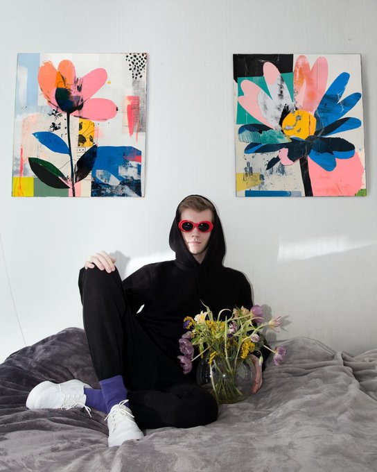 A person wearing a black hoodie, red sunglasses, purple socks, and white sneakers sits on a gray blanket holding a vase of flowers, with two colorful abstract floral paintings hanging on the white wall behind them.