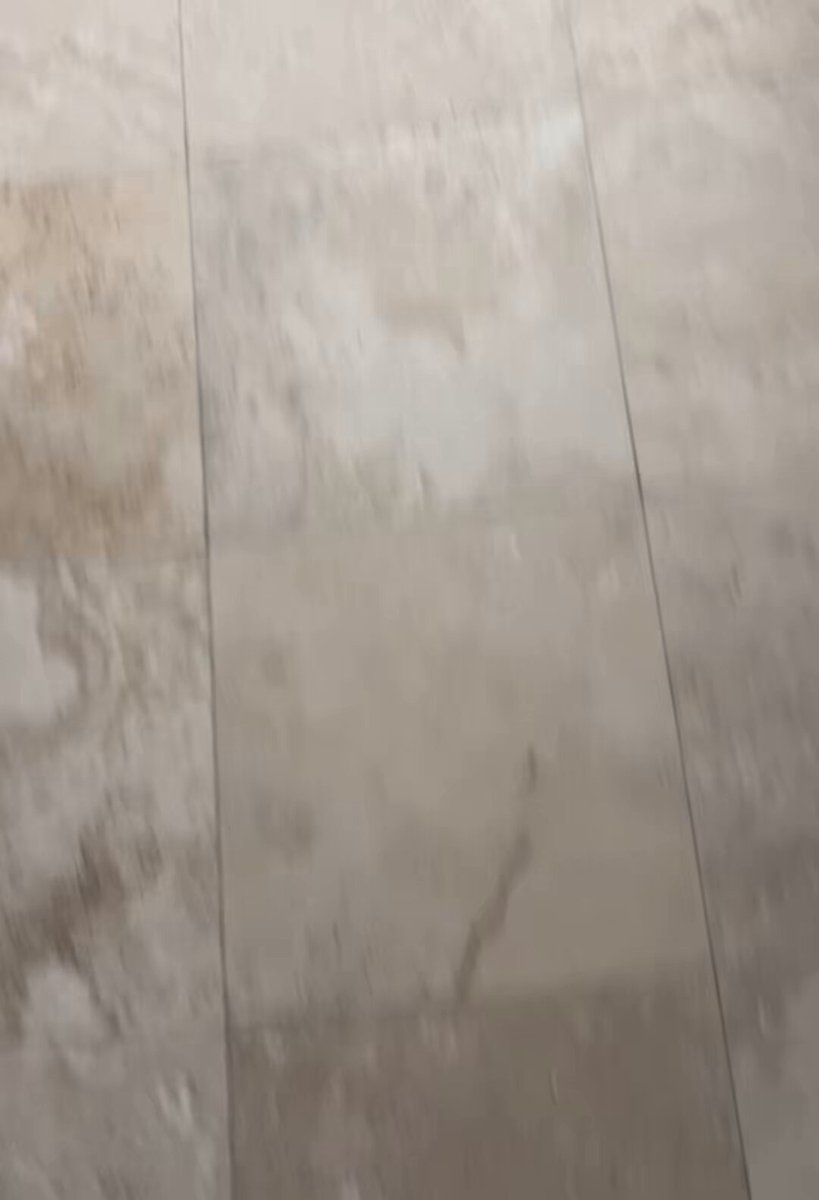 The image shows a close-up of a floor with a tile pattern, possibly marble or beige stone tiles. The perspective suggests that someone took the photo from below, looking up at the floor. In the background, there are two people present - one person is panicking and the other appears to be laughing. This scene could be related to an event where XRP was being discussed, such as a market drop or a cryptocurrency conference. The individuals reacting differently to this situation might indicate varying levels of understanding or interest in the subject matter.