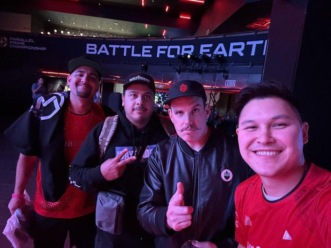 Four men pose for a photo in front of a 'BATTLE FOR EARTH' and 'PARALLEL PRIME CHAMPIONSHIP' sign at what appears to be a gaming/esports event, with three wearing red shirts and giving thumbs up.