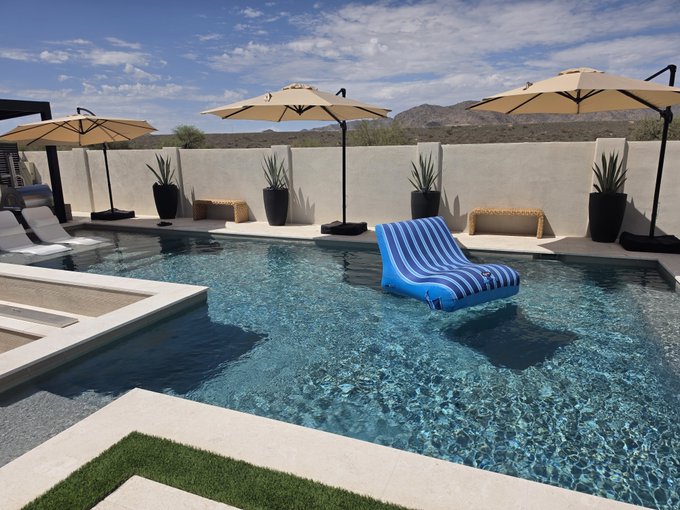 An infinity-edge swimming pool features a floating blue and white striped lounge chair, surrounded by tan umbrellas, potted plants, and desert landscape under a partly cloudy sky.