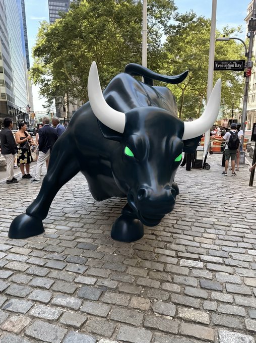 A black bull statue with white horns and glowing green eyes positioned on a cobblestone street, with pedestrians and a sign reading 'Evacuation Day' visible in the background.