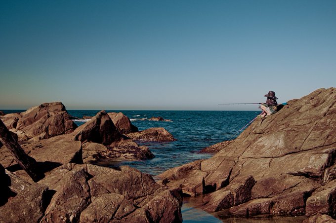 A person sits on rocky coastal terrain fishing in the ocean under a clear blue sky, with rugged brown rocks in the foreground.