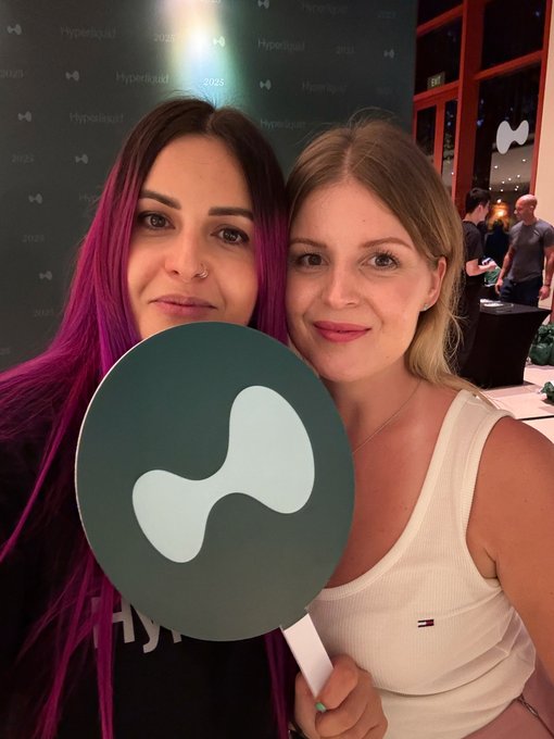 The image features two women standing next to each other, both holding up a large sign with a white circle design. The sign has the word "Hyperliquid" written on it. They are posing for a photo together, possibly at an event or gathering related to Hyperliquid.