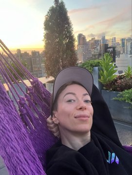 A selfie of a young woman wearing a cap and black hoodie, relaxing in a purple macramé hammock with a city skyline at sunset in the background.