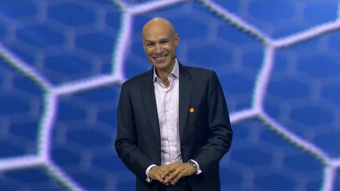 A bald man in a navy suit and light purple shirt smiles broadly while standing in front of a blue hexagonal patterned backdrop.