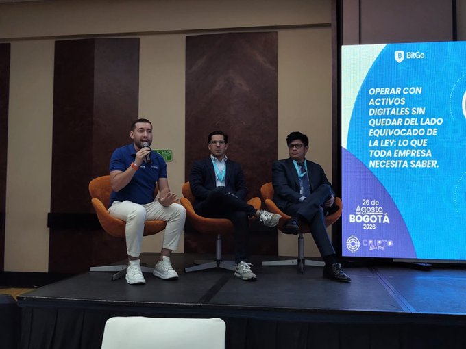 Three men seated on a stage at a panel discussion, with a screen displaying "OPERAR CON ACTIVOS DIGITALES SIN QUEDAR DEL LADO EQUIVOCADO DE LA LEY" and event details for "BOGOTÁ 2028."