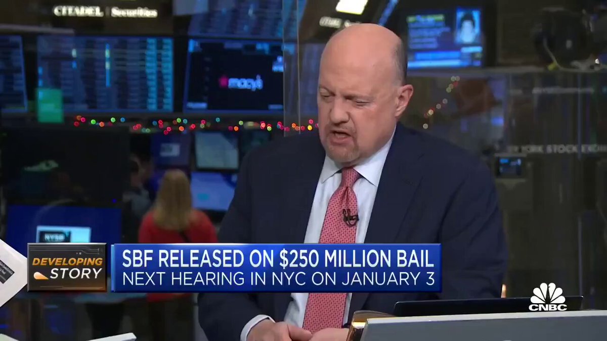 The image shows a man wearing a suit, sitting at a desk with a laptop computer open in front of him. The background features multiple screens displaying stock market information, including the names "S&P 500" and "IBM". The text on the screen reads "Just In: Jim Cammer just said his pro-bitcoin stance", indicating that Jim Cammer has made a statement about Bitcoin or cryptocurrency.
