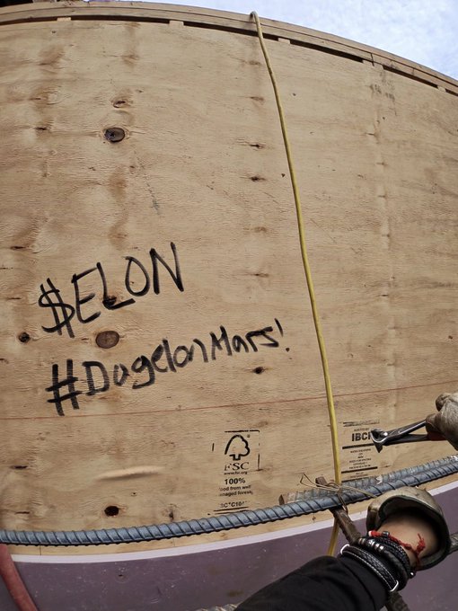 A wooden crate marked '#ELON' and '#DogelonMars!' being held by a worker wearing a glove, secured with a yellow strap.