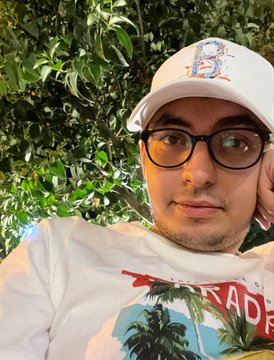 A man wearing a white cap and glasses poses for a close-up selfie against a leafy green background, sporting a white graphic tee with "PARADISE" visible. The image aligns with "time to look real" by showcasing a candid, unfiltered moment where he appears to be presenting himself authentically in a natural outdoor setting.
