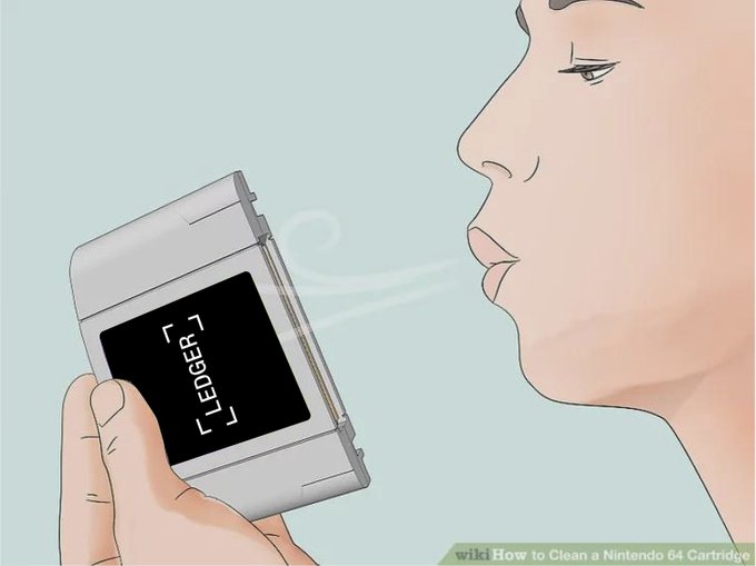 An illustration from a wikiHow article titled 'How to Clean a Nintendo 64 Cartridge' shows a person blowing breath onto a gray N64 game cartridge labeled 'LEDGER.'