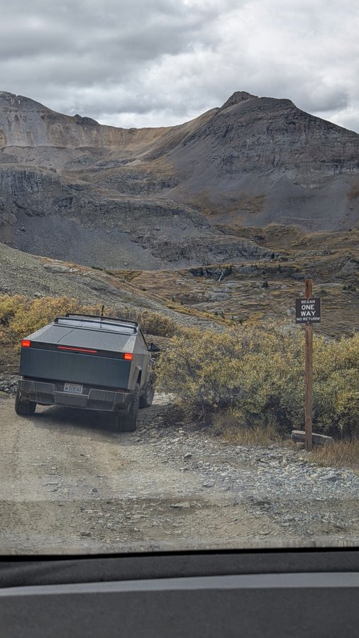 A Tesla Cybertruck parked on a gravel road in a mountainous landscape, next to a wooden post bearing a 'ONE WAY' sign.