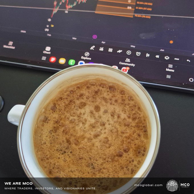 The image features a cup of coffee sitting on top of a desk next to a computer screen displaying stock market data. The coffee appears to be frothy, suggesting that it has been freshly prepared or recently poured. The presence of the computer screen with stock market information indicates that someone is likely working in finance or analyzing financial trends. The image captures a moment where work and personal interests intersect, as evidenced by the cup of coffee and the computer screen displaying stock market data.