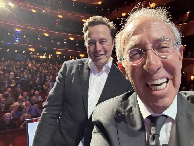 A selfie-style photo shows Elon Musk and Ron Baron smiling together inside a packed theater, with Musk wearing a dark suit and Baron in the foreground wearing glasses and a tie, capturing a candid moment after Baron's interview that the accompanying Korean post humorously reimagines as a fictional conversation where a weary investor pleads with Musk for a stock price rebound.