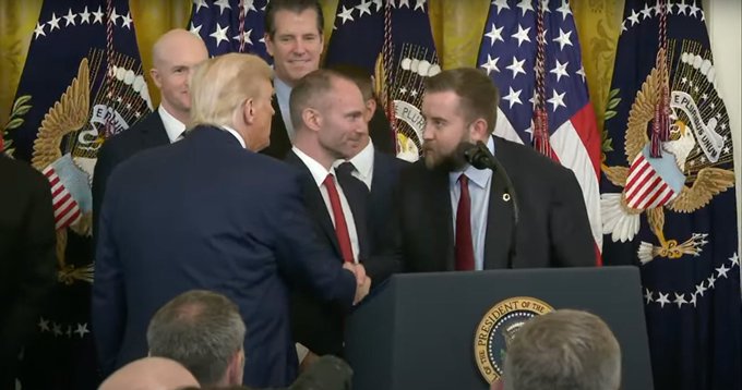 President Donald Trump shakes hands with a bearded man in a suit at a podium bearing the Presidential Seal, while other officials, including a smiling man who appears to be Senator Mark Warner, stand behind them in front of American flags. The image ties to the post by depicting the political setting in which the Clarity Act—a bill viewed by crypto supporters as supportive of Chainlink ($LINK)—was signed or celebrated.
