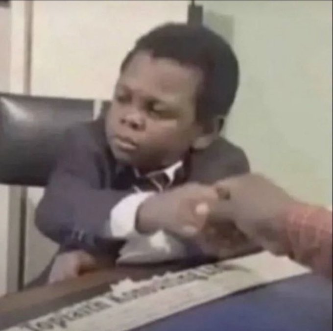 A low-quality image of a young boy sitting at a table in a suit, pointing at something off-screen with a serious expression.
