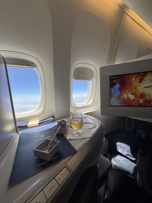 An airplane window seat view showing two oval windows with sky and clouds visible, alongside a tray with a drink, utensils, and a screen displaying cartoon characters.