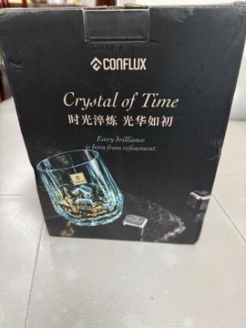 A black Conflux branded gift box labeled 'Crystal of Time' with Chinese text '时光淬炼 光华如初' and the tagline 'Every brilliance born from refinement.', containing a whiskey glass with a gold ship inside.