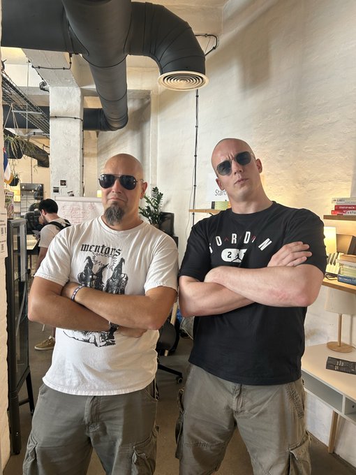 Two bald men with goatees pose indoors with arms crossed; one wears a white 'mentors' t-shirt and sunglasses, while the other wears a black 'ORION' t-shirt and sunglasses, in what appears to be an office or workspace.