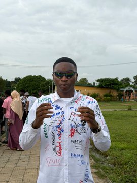 A young man wearing sunglasses and a white shirt covered in colorful signatures poses outdoors on a paved area with trees and people in the background.