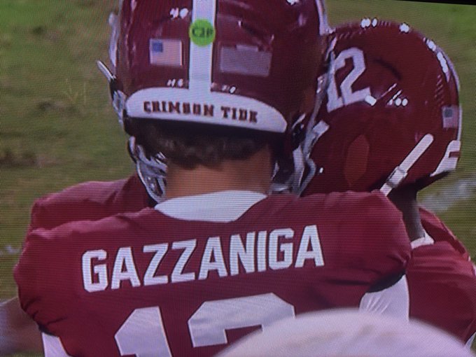 Two Alabama Crimson Tide football players in crimson helmets embrace on the field, with the jersey of one clearly reading 'GAZZANIGA' and the helmet displaying 'CRIMSON TIDE'.