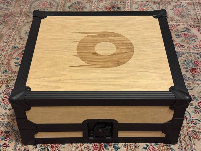 A luxurious wooden and metal poker case featuring an engraved comet/celestial logo sits on a floral patterned rug, presented as a custom one-of-a-kind set from Rollbit. The case showcases premium craftsmanship with its reinforced corners and latched clasp, matching the post's celebration of receiving this exclusive Rollbit collaboration.