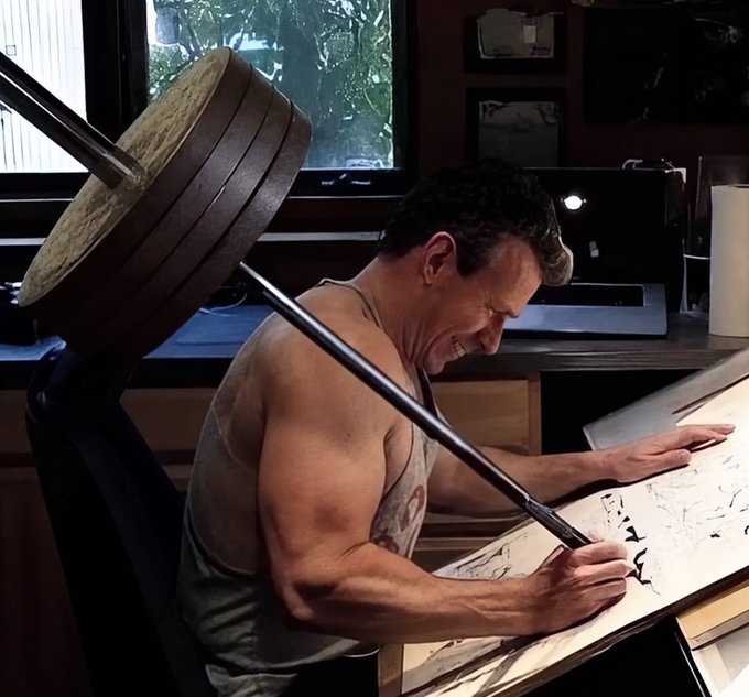 A muscular man in a grey tank top sits at a desk, intensely drawing in a sketchbook under a large studio lamp.