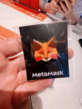 A hand holds up a clear plastic pouch featuring a fox logo and 'MetaMask' branding, likely a promotional item from a crypto/web3 event.