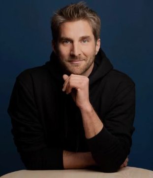 A portrait of a smiling man with brown hair and a beard, wearing a black hoodie, seated at a table against a blue background with his chin resting on his hand.