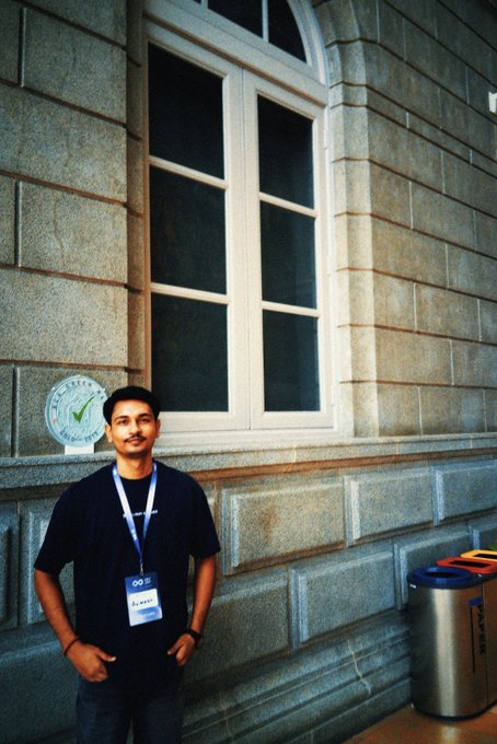 A young man wearing glasses, a navy t-shirt, and a lanyard stands smiling in front of a stone building with large windows and trash bins nearby.