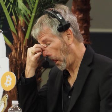 The image depicts a man wearing a headset, leaning over a table with his head down while looking at something off screen. The scene also includes bottles of water placed near him. In the background, there's a palm tree, and a keyboard is visible on one side. 

In the provided text, MicroStrategy has suffered massive losses with assets evaporating to a combined value of $16 billion. Tom Lee Bitmine, another company in the same industry as Microstrategy, has held 2 Bitcoin Bitcoins for an average cost of $75,699 and is suffering a loss of $7.3 billion.