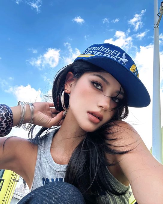 A young woman wearing a blue cap with "Hollister" text poses for a selfie against a bright blue sky, accessorized with hoop earrings and silver bracelets. The casual, sunlit shot matches the friendly "Gm fam" greeting, giving off a relaxed, social-media-ready vibe.