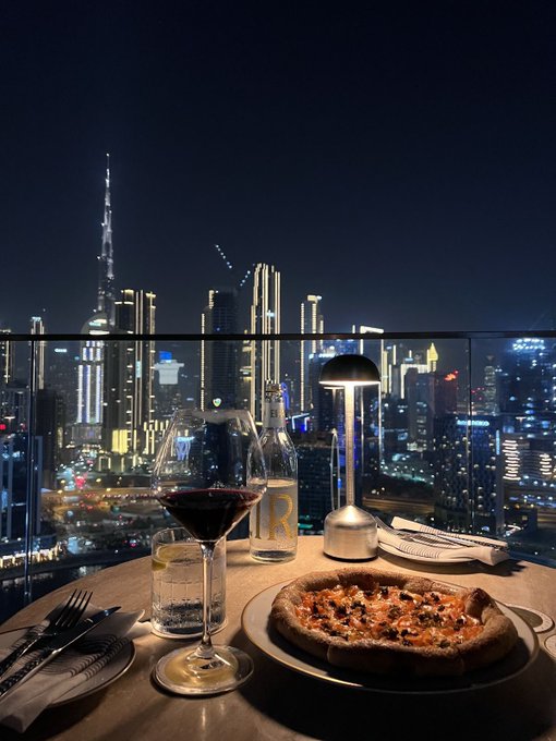 The image features a table with a pizza placed on top of a plate, accompanied by wine glasses filled with red wine. The scene takes place at night, with a city skyline visible through a glass window or balcony. The focus of the image is on the delicious meal and the beautiful view outside.