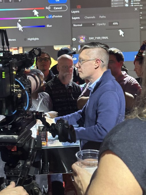 Behind-the-scenes photo of a man wearing glasses and a blue jacket operating a professional film camera on a set with crew members in the background.
