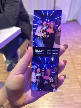 The image shows a person holding up a photo of themselves at an event, likely a summit or conference. The photo captures four people posing for a picture together. The background appears to be a room with a wooden floor and white walls. There is also a chair visible in the scene. The caption on the post reads "Solana Summit Germany is a banger. Will share a video soon with the highlights." This suggests that the person holding up the photo might have attended or will attend an event at Solana Summit Germany, and they want to share their experience by taking a picture of themselves and their friends.