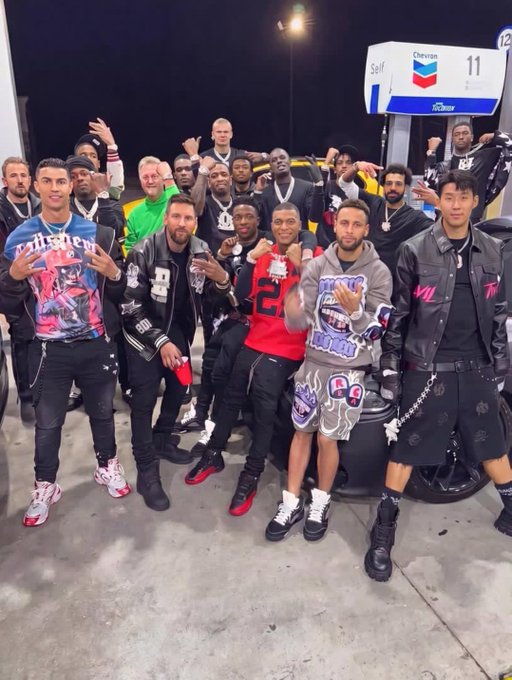 A group of men pose for a photo at a Chevron gas station, standing near pump number 11. Several are wearing black jackets with patches, suggesting they may be part of a club or group gathering.