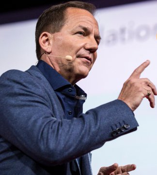 A man in a blue blazer with a headset microphone gestures while speaking on stage.