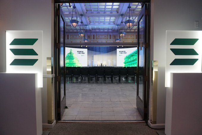 An event hall entrance with double doors flanked by illuminated green arrow signs, leading into a room with rows of black chairs and a stage with screens reading 'INNOVATION HALL STREET'.