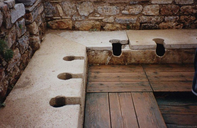 Ancient Roman-style stone latrines with keyhole-shaped openings built into a wall, with a wooden floor below.
